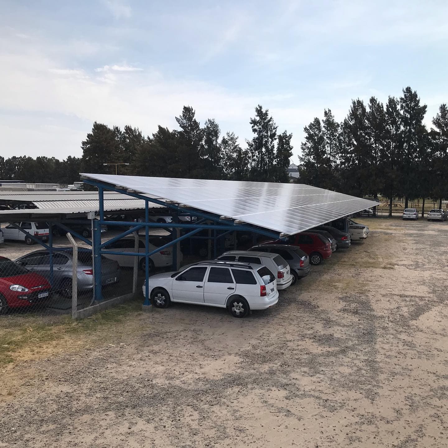 Estructura azul con un techo de paneles que da sombra a una fila de autos estacionados.